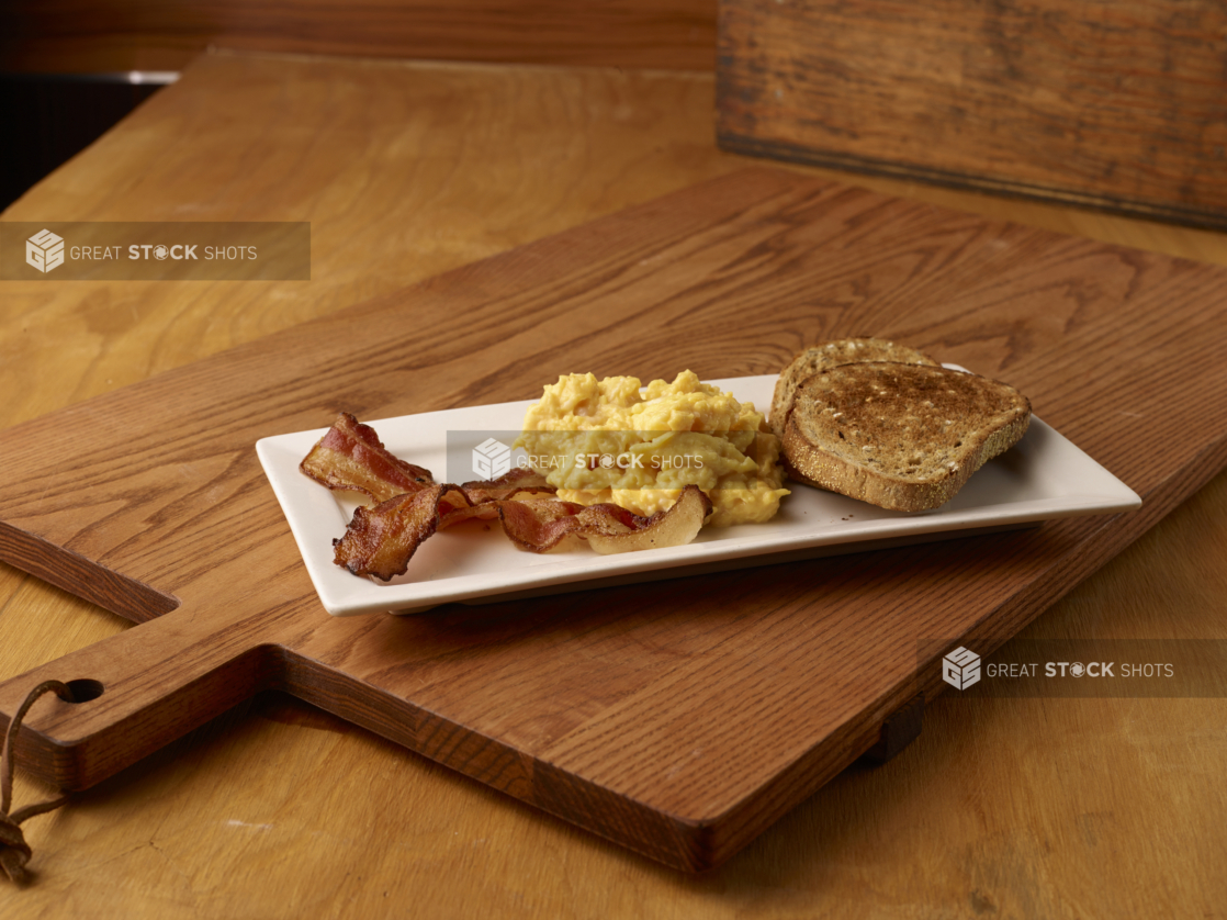 Sliced Toasted Rye Bread with Scrambled Bread and Bacon Strips on a Rectangular White Platter Dish on a Wooden Cutting Board