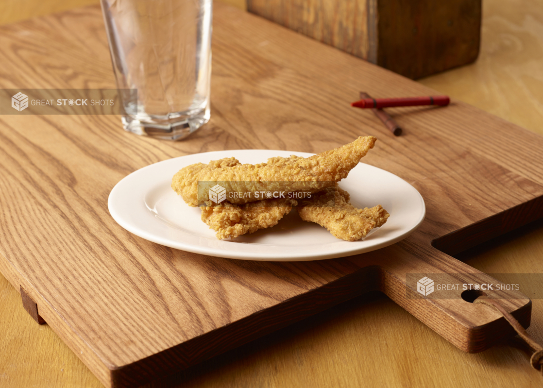 Deep Fried Breaded Chicken Tender Strips on a Round White Plate with a Glass of Water on a Wooden Cutting Board with Crayons for Kids