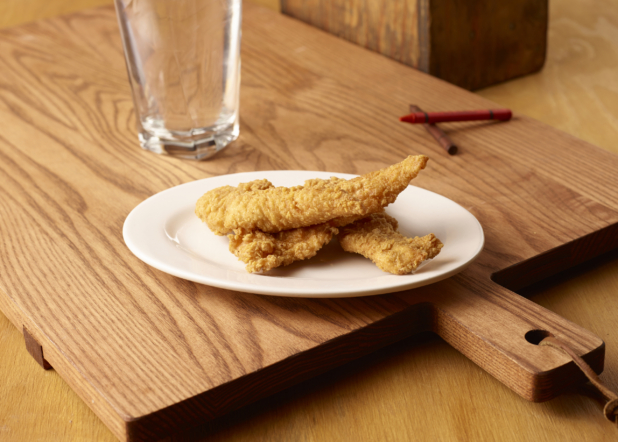 Deep Fried Breaded Chicken Tender Strips on a Round White Plate with a Glass of Water on a Wooden Cutting Board with Crayons for Kids