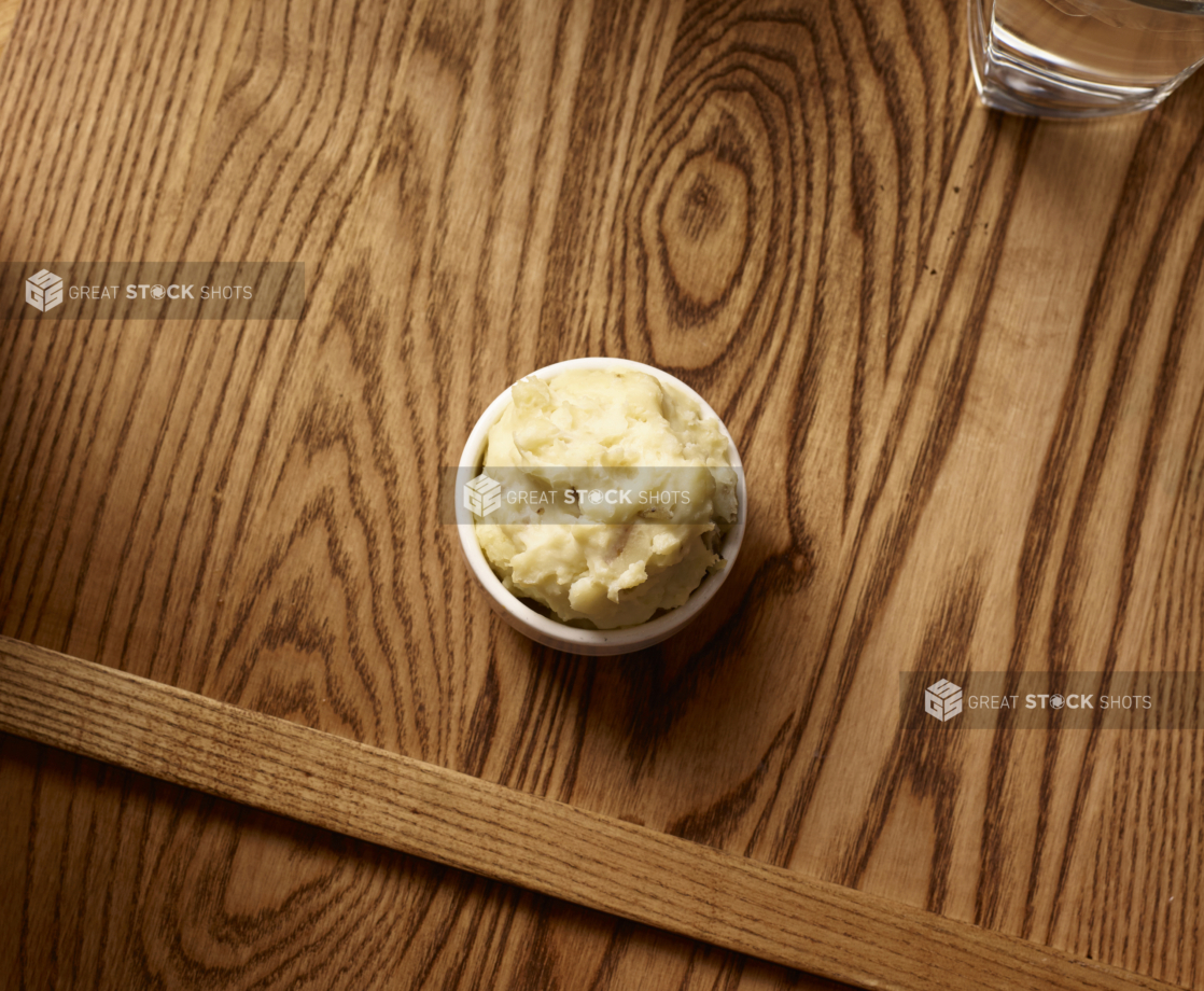 Overhead View of a Small White Ceramic Dish of Kid-Sized Mashed Potatoes Side Dish on a Wooden Cutting Board in an Indoor Setting