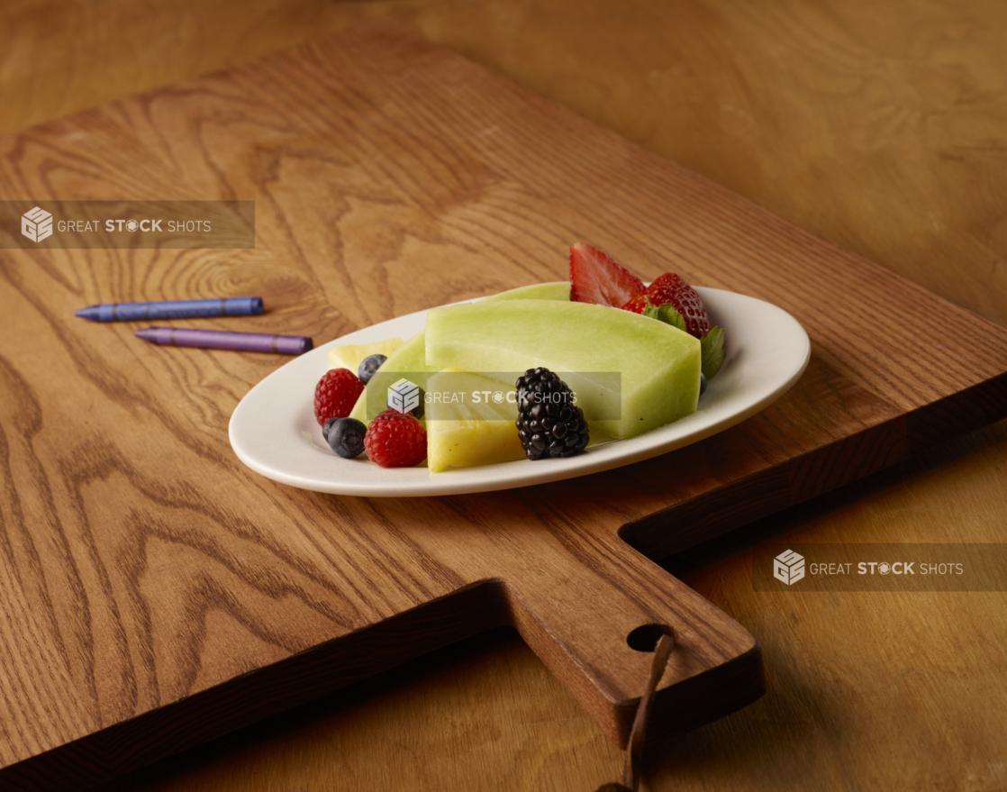 Round White Ceramic Plate of Cut Honeydew Melon, Pineapple, Blackberries, Blueberries, Raspberries and Strawberries on a Wooden Cutting Board in an Indoor Setting