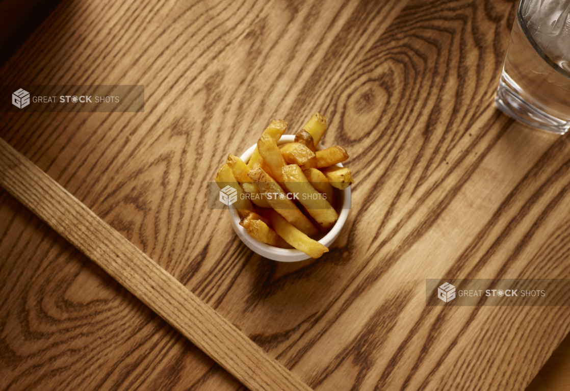 Overhead View of a Small Kids Size Thick Cut French Fries in a Round White Bowl on a Wooden Cutting Board in an Indoor Setting