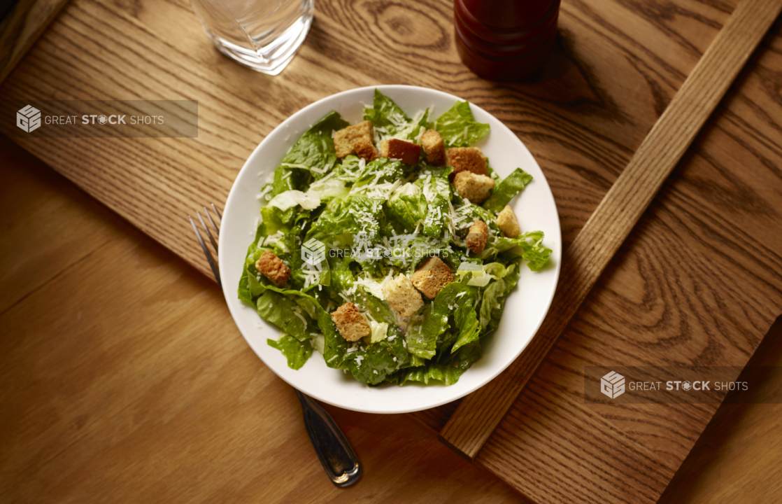 Overhead View of a Caesar Salad in a Round White Ceramic Dish on a Wooden Cutting Board in an Indoor Setting