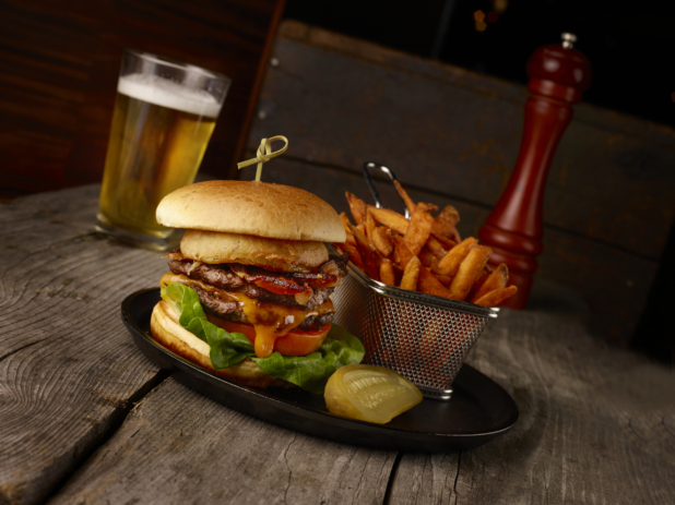 Gourmet Double Bacon Cheeseburger with a Side of Sweet Potato Fries and a Glass of Draught Beer on a Rustic Wooden Table in an Indoor Setting