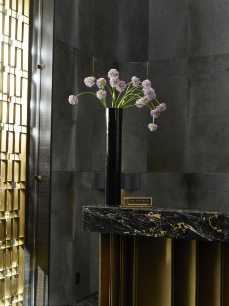 A Tall Black Cylindrical Vase of Light Purple Allium Flowers on a Black Marble Reception Desk in an Indoor Setting
