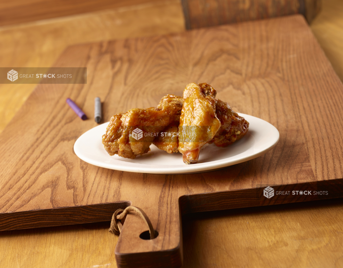 Round White Ceramic Dish of Kid-Sized BBQ Chicken Wings on a Wooden Cutting Board in an Indoor Setting