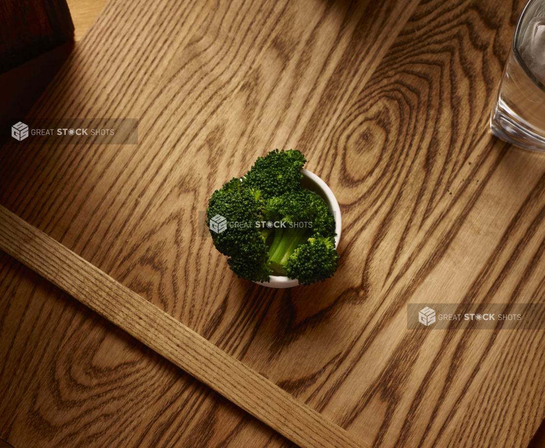 Overhead View of a Small White Ceramic Dish of Kid-Sized Steamed Broccoli Side Dish on a Wooden Cutting Board in an Indoor Setting