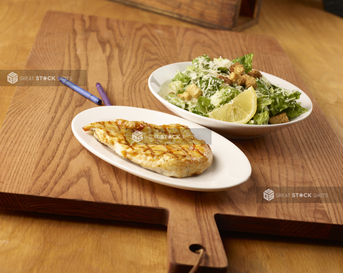 Kids Meal of Grilled BBQ Chicken Breast on a Round White Dish and a Small Side of Caesar Salad on a Wooden Cutting Board in an Indoor Setting