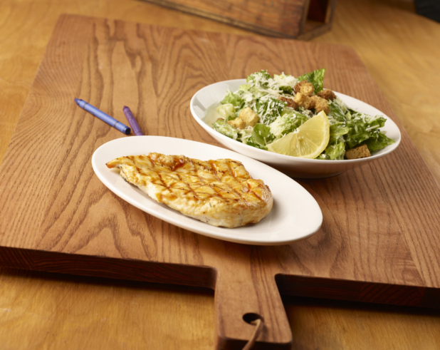 Kids Meal of Grilled BBQ Chicken Breast on a Round White Dish and a Small Side of Caesar Salad on a Wooden Cutting Board in an Indoor Setting