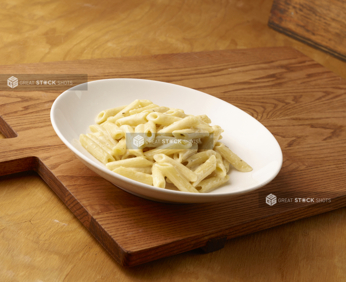 Kids Penne Alfredo Pasta in a Large White Ceramic Bowl on a Wooden Cutting Board in an Indoor Setting