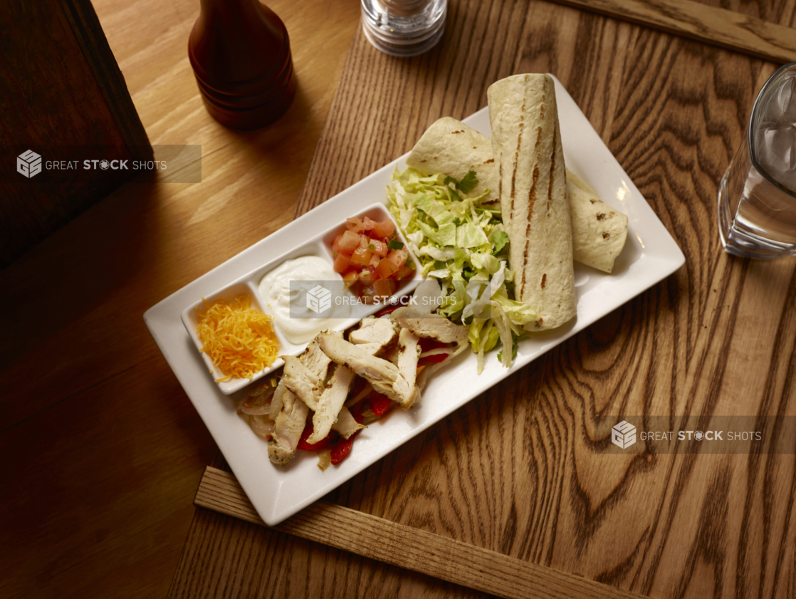 Overhead View of a Kids Grilled Chicken Fajita Platter on a Rectangular White Ceramic Dish with Fresh and Cooked Ingredients and Toppings on a Wooden Cutting Board in an Indoor Setting