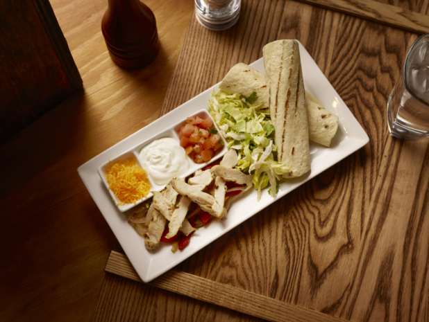 Overhead View of a Kids Grilled Chicken Fajita Platter on a Rectangular White Ceramic Dish with Fresh and Cooked Ingredients and Toppings on a Wooden Cutting Board in an Indoor Setting