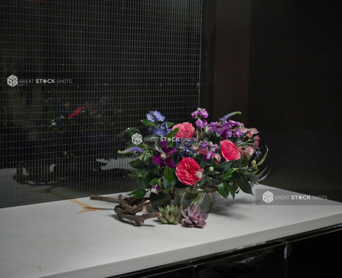 Floral Centrepiece of Deep Pink Garden Roses, Purple Clematis, Blue and Magenta Dephiniums in a Large Glass Vase with Green and Ruby-Kissed Echeveria Succulents