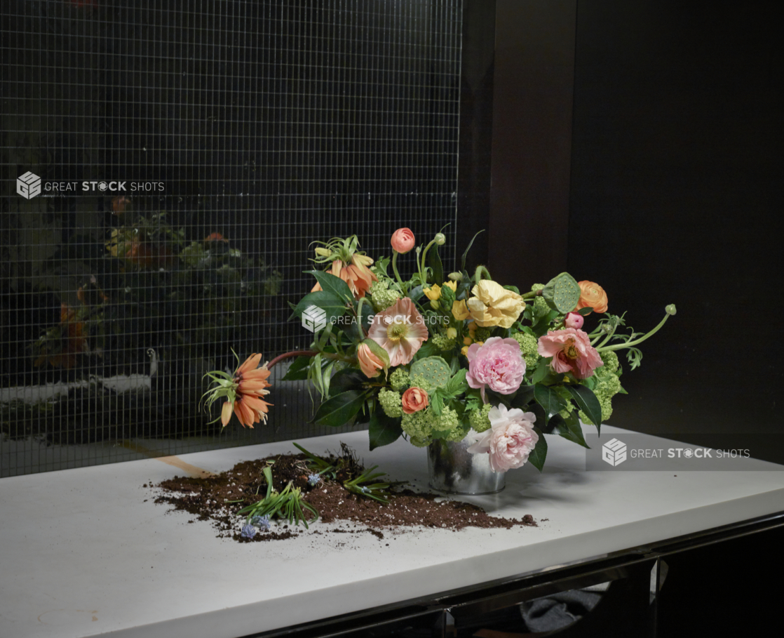 Floral Centrepiece of Light Pink Peonies, Mini White Hydrangeas, Orange Fritillaria and Lotus Seed Pods in Preparation Stages with Pot Soil on a Table