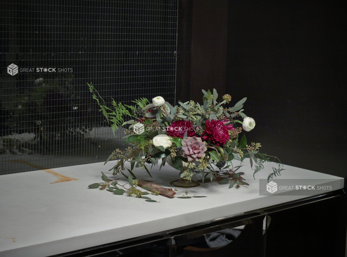 Flower arrangement of red chrysanthemums and white ranunculus on a white marble countertop in front of a mirrored mosaic