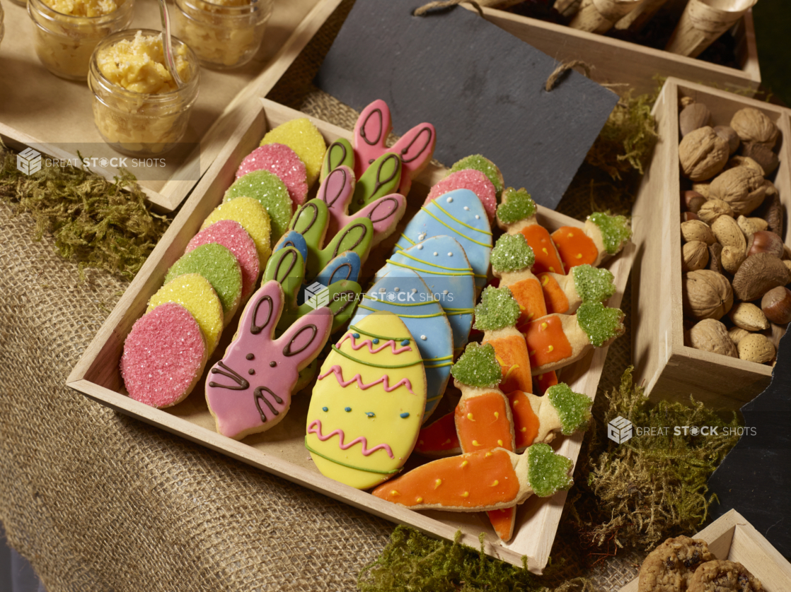 Brightly decorated Easter cookies in a wood serving tray surrounded by various holiday treats on a burlap-covered table