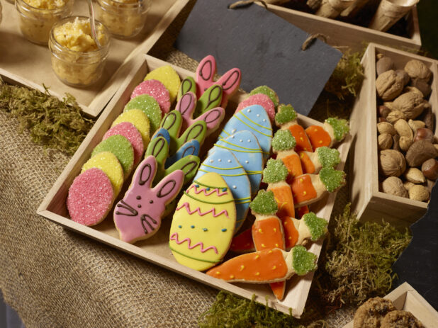 Brightly decorated Easter cookies in a wood serving tray surrounded by various holiday treats on a burlap-covered table