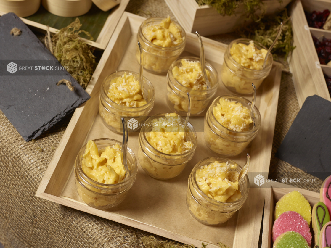 Mac and Cheese in Glass Mason jars with Small Spoons on a Wood Catering Tray on a Burlap Surface in an Indoor Setting