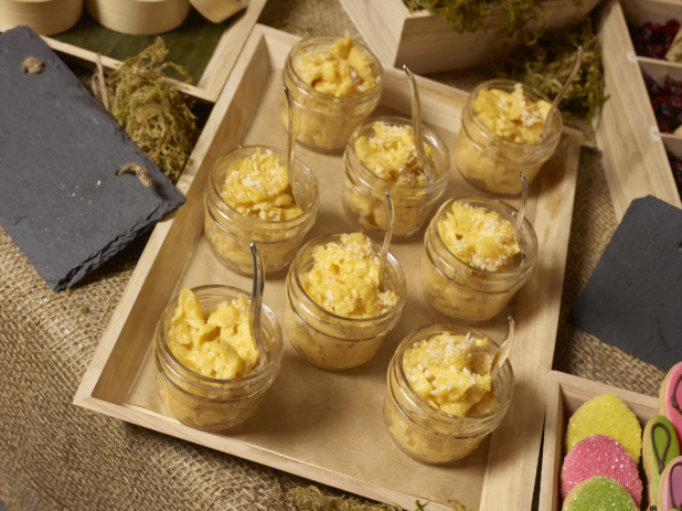 Mac and Cheese in Glass Mason jars with Small Spoons on a Wood Catering Tray on a Burlap Surface in an Indoor Setting