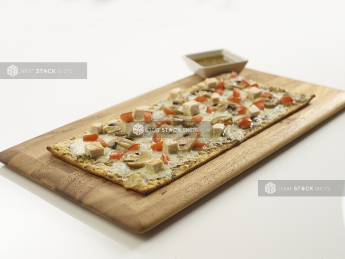 Rectangular flatbread with melted cheese, chicken cubes, mushrooms, and tomatoes on a wooden board, white background