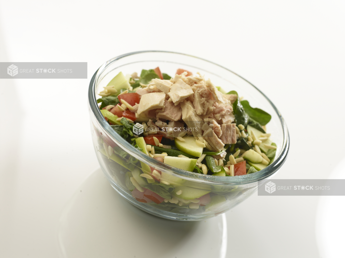 Spinach salad with tuna, cucumbers, tomato, and slivered almonds in a clear glass bowl on a reflective white surface