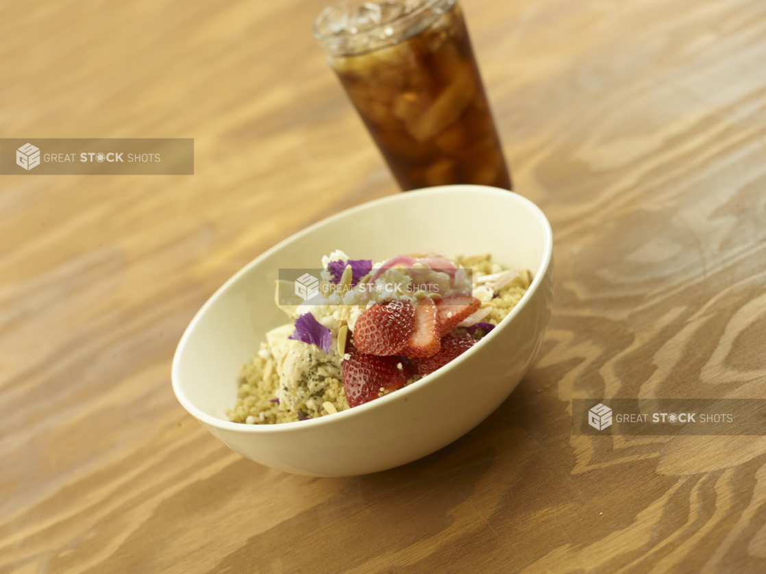 A Breakfast Bowl of Quinoa, Sliced Strawberries, Sliced Almonds and Yogurt with a Glass of Diet Coke and Ice on a Wooden Table in an Indoor Setting