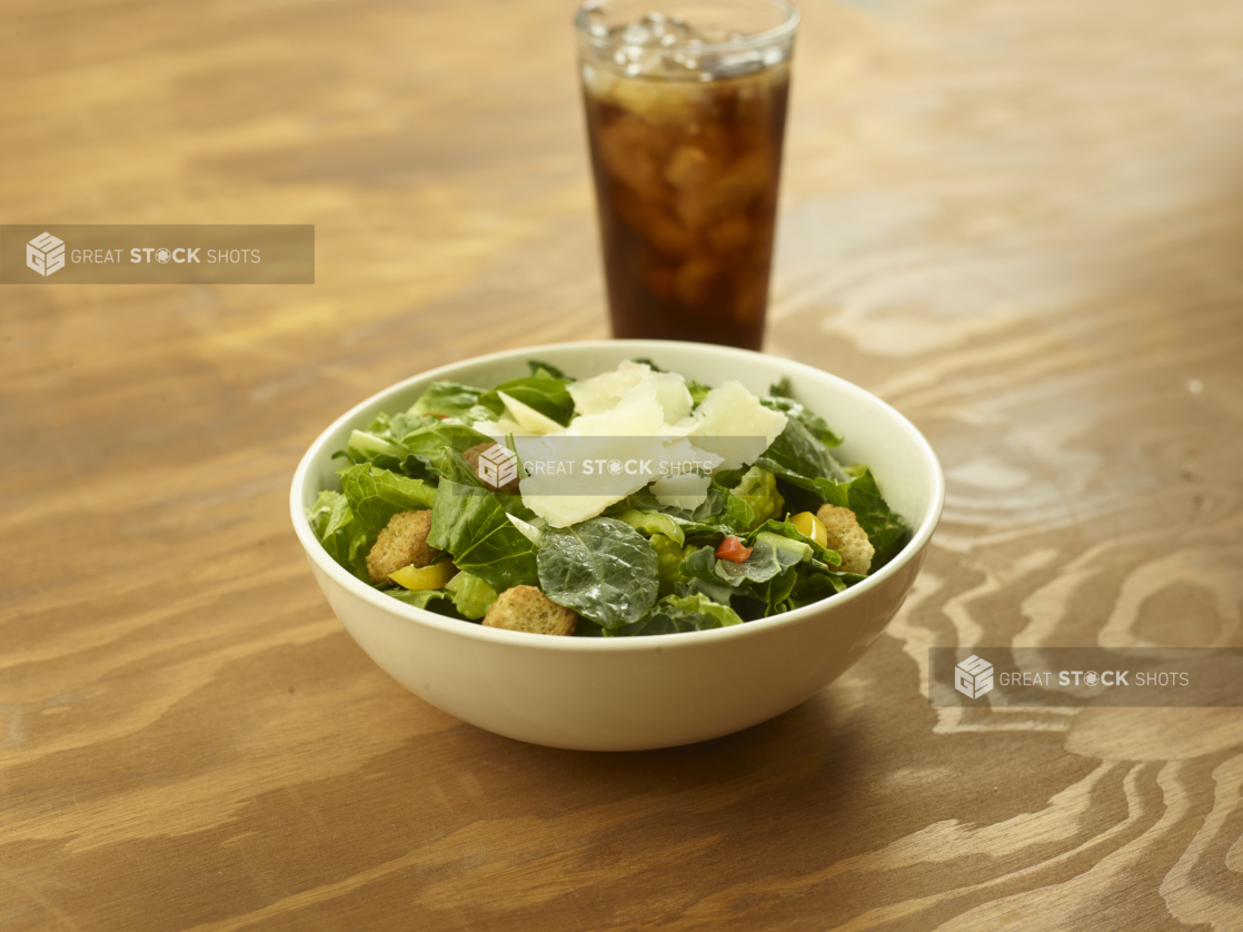 A Bowl of Baby Spinach and Romaine Caesar Salad with Colourful Peppers, Croutons and Shaved Parmesan and a Glass of Diet Coke and Ice on a Wooden Table in an Indoor Setting