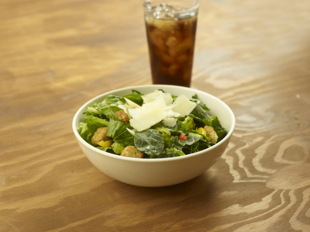 A Bowl of Baby Spinach and Romaine Caesar Salad with Colourful Peppers, Croutons and Shaved Parmesan and a Glass of Diet Coke and Ice on a Wooden Table in an Indoor Setting