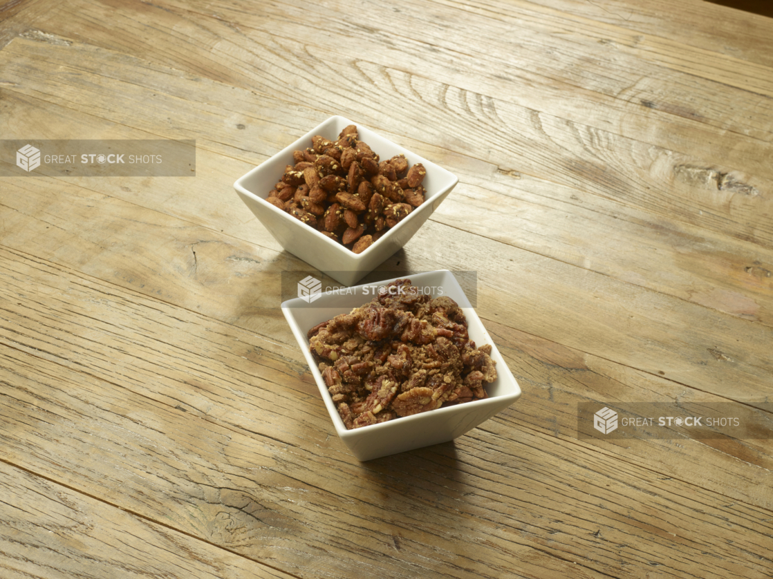 Two White Square Bowls Filled with Smoked Pecans and Almonds and Assorted Nuts on a Wooden Table