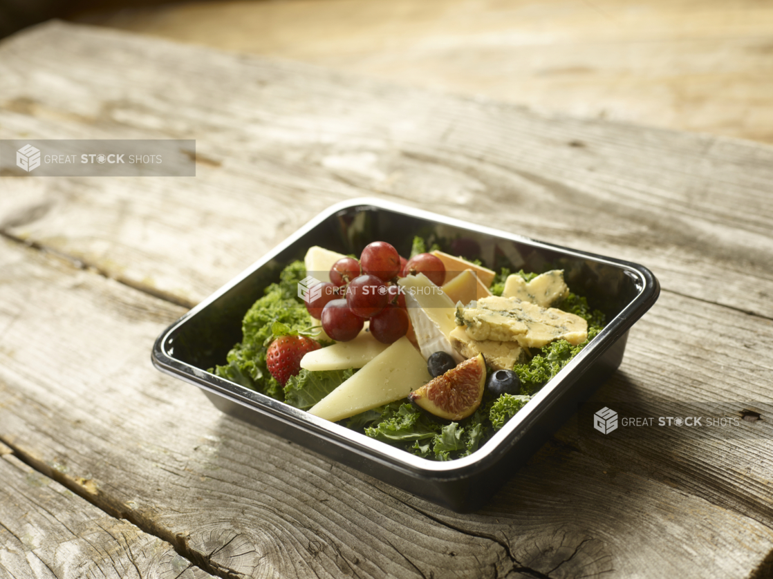 A Healthy Snack of Assorted Cheese Slices, Fruits on a Bed of Lettuce in a Black Take-Out Container on a Wooden Table