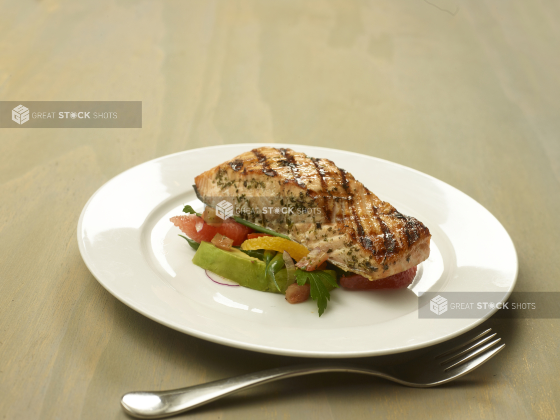 Herb-Crusted Grilled Salmon Fillet on a Bed of Ruby Red Grapefruits and Avocado on a Round White Ceramic Dish on a Wooden Surface in an Indoor Setting