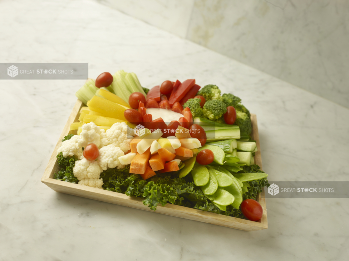 Vegetable Crudité Platter Featuring Assorted Raw Vegetables and Veggie Sticks on a Bed of Kale with a Red Pepper Tulip Cup of Creamy Dipping Sauce on a Square Wood Serving Tray on a Marble Surface