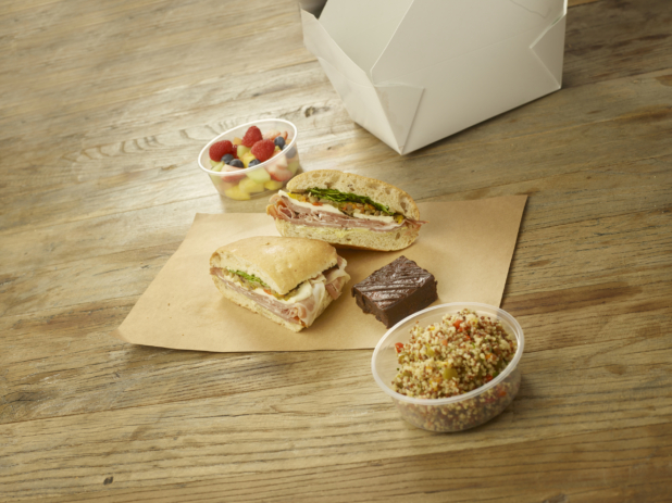 A Boxed Lunch of an Italian Muffuletta Sandwich, Quinoa Salad, Fresh Cut Fruit and a Chocolate Brownie with a White Cardboard Take-Out Container on a Rustic Wooden Surface