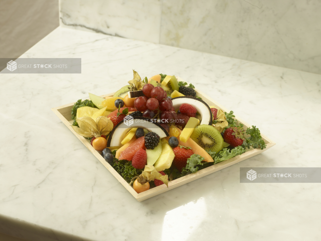 Assorted Sliced and Whole Fruits on a Bed of Romaine Lettuce in a Square Wood Tray on a Marble Surface - for Catering