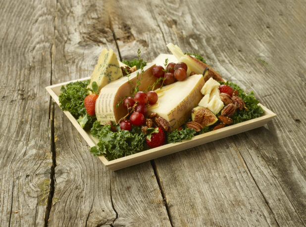 Close Up of Assorted Sliced and Cut Cheeses with an Assortment of Fruit and Nuts on a bed of Kale Leaves in a Square Wooden Tray on an Aged Wooden Table