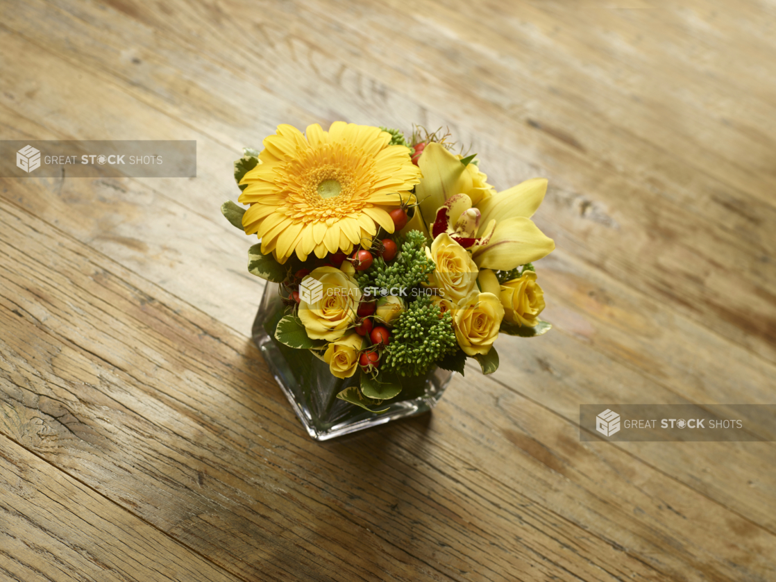 Glass Cube Vase with Yellow Rose, Lily, Daisy Flowers and Green Sedum and Hypericum Berries on a Rustic Wooden Surface in an Indoor Setting