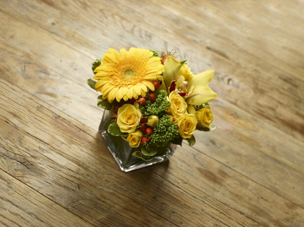 Glass Cube Vase with Yellow Rose, Lily, Daisy Flowers and Green Sedum and Hypericum Berries on a Rustic Wooden Surface in an Indoor Setting