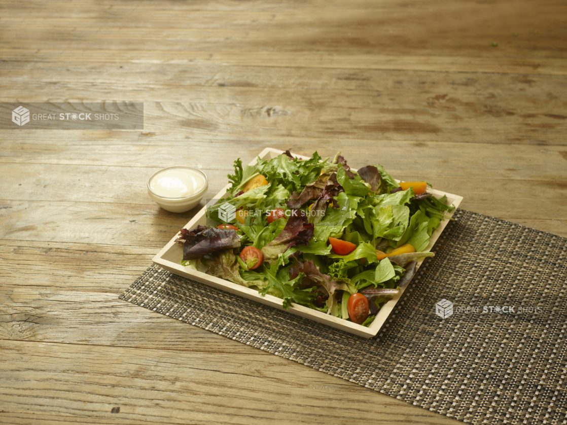 A Square Wood Serving Tray with Spring Mix, Cherry and Grape Tomato Salad on a Woven Placemat on a Rustic Wooden Surface with a Side Dish of Creamy Salad Dressing in an Indoor Setting