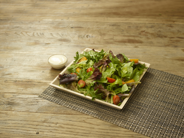A Square Wood Serving Tray with Spring Mix, Cherry and Grape Tomato Salad on a Woven Placemat on a Rustic Wooden Surface with a Side Dish of Creamy Salad Dressing in an Indoor Setting