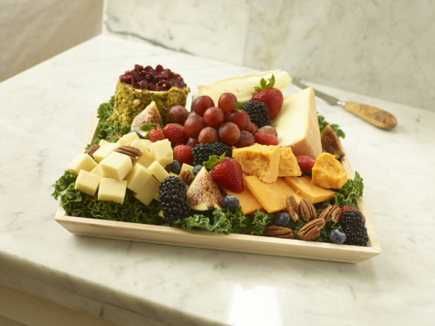 Assorted Sliced and Cut Cheeses with an Assortment of Fruit and Pistachio-Crusted Goat Cheese on a bed of Kale Leaves in a Square Wooden Tray on a Marble Counter
