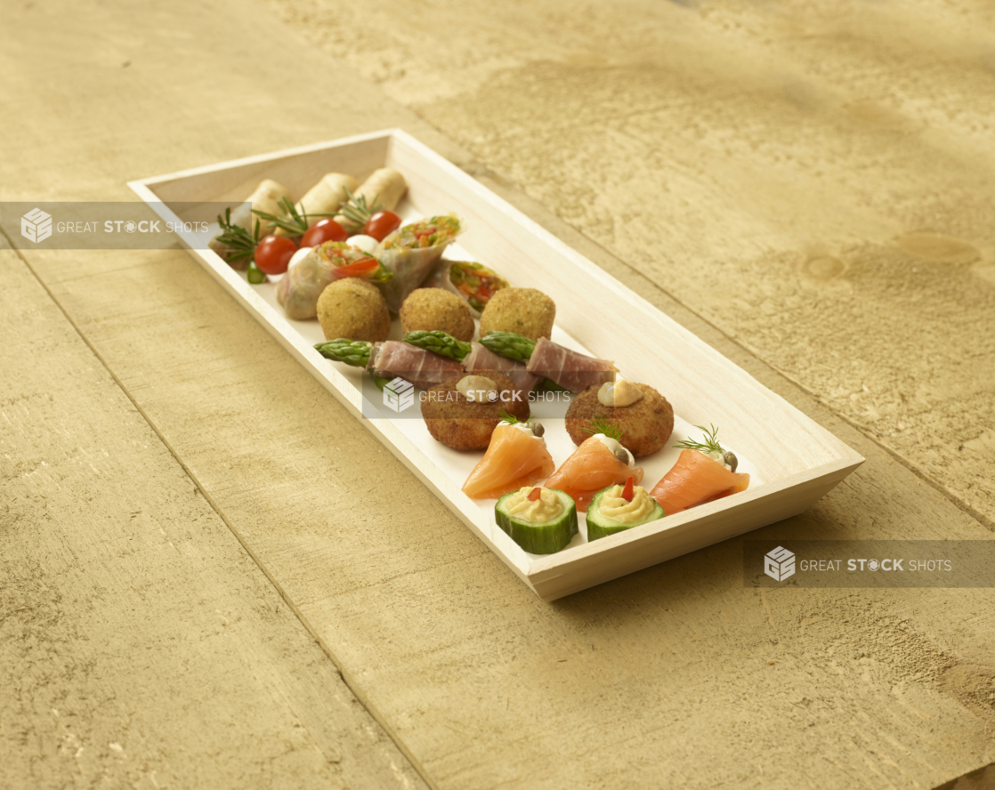 A Narrow Rectangular Wood Serving Tray with Assorted Passed Appetizers (Smoked Salmon Rolls, Croquettes, Spring Rolls, Cucumber Bites, Caprese Skewers) for Catering on an Untreated Wood Surface