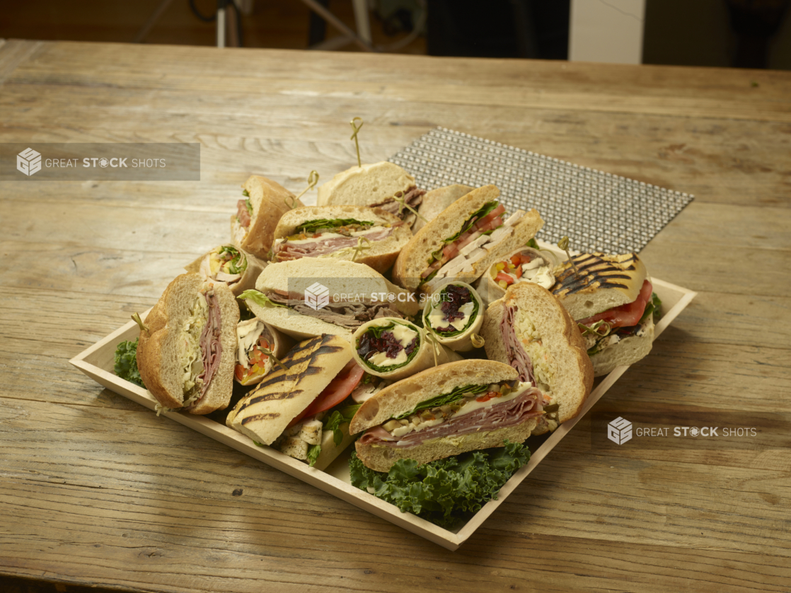 A Square Catering Tray with an Assortment of Cold Sandwiches and Wraps Cut in Half on a Bed of Kale Leaves on a Wooden Surface