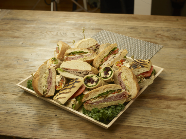A Square Catering Tray with an Assortment of Cold Sandwiches and Wraps Cut in Half on a Bed of Kale Leaves on a Wooden Surface