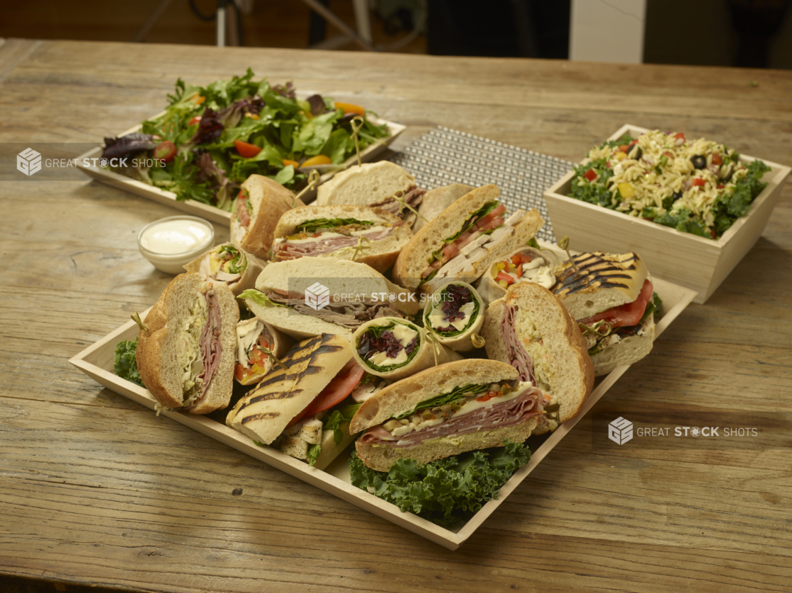 A Lunch Catering Package with an Assorted Sandwich Platter, Spring Mix Salad and Orzo Pasta Salad in Wood Serving Trays on a Wooden Table