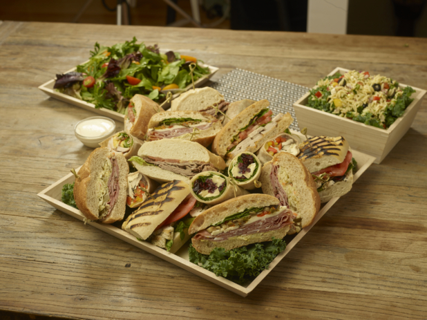 A Lunch Catering Package with an Assorted Sandwich Platter, Spring Mix Salad and Orzo Pasta Salad in Wood Serving Trays on a Wooden Table