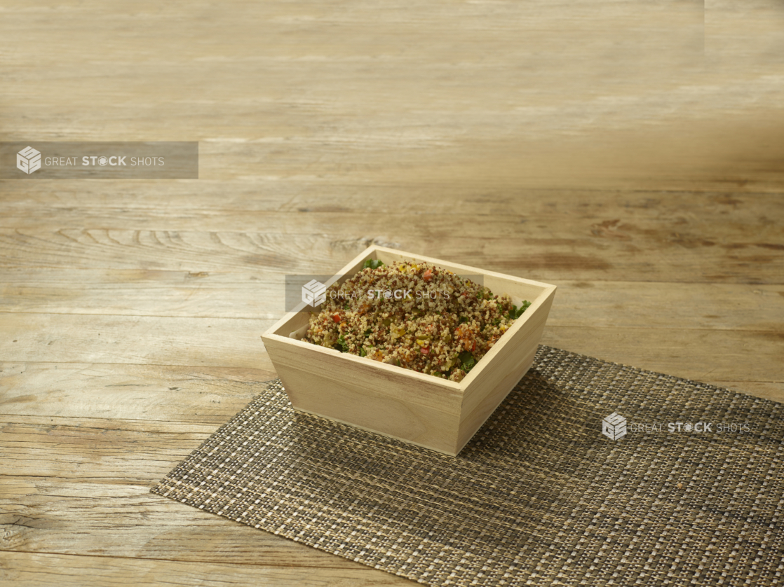 A Small Deep Square Wood Serving Tray of Quinoa Salad on a Woven Placemat on a Rustic Wood Surface in an Indoor Setting