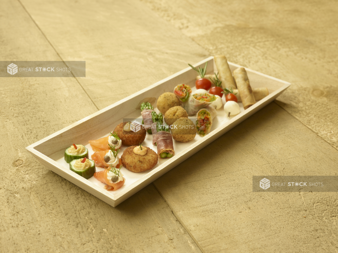 A Narrow Rectangular Wood Serving Tray with Assorted Passed Appetizers (Smoked Salmon Rolls, Croquettes, Spring Rolls, Cucumber Bites, Caprese Skewers) for Catering on an Untreated Wood Surface - Variation