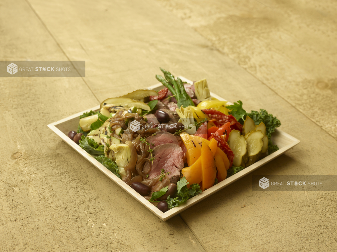 Square Wood Serving Tray with Pork Tenderloin and Assorted Roasted Vegetables and Pickles on a Bed of Kale on an Untreated Wood Surface