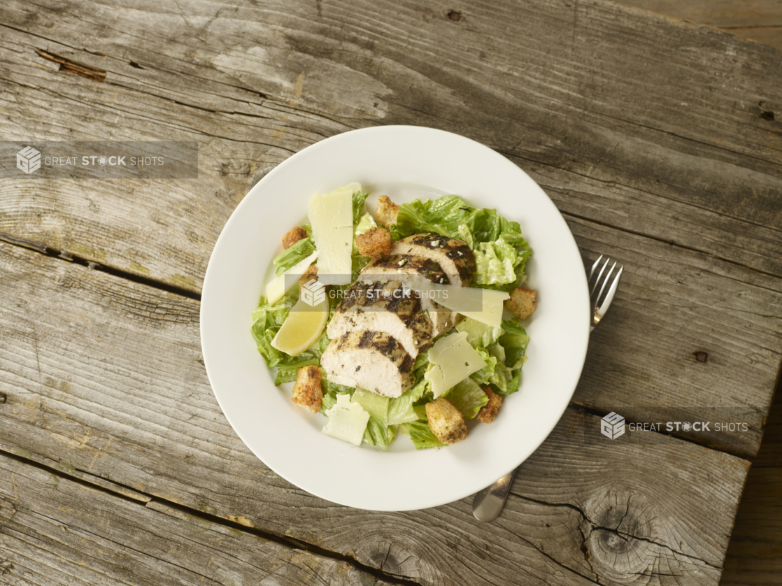 Overhead View of a Grilled Chicken Caesar salad with Shaved Parmesan Cheese, Croutons and a Lemon Wedge on a Round White Ceramic Dish on a Rustic Wooden Surface