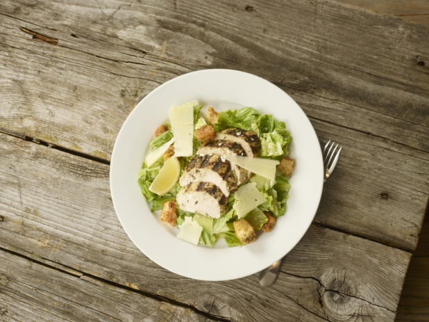 Overhead View of a Grilled Chicken Caesar salad with Shaved Parmesan Cheese, Croutons and a Lemon Wedge on a Round White Ceramic Dish on a Rustic Wooden Surface